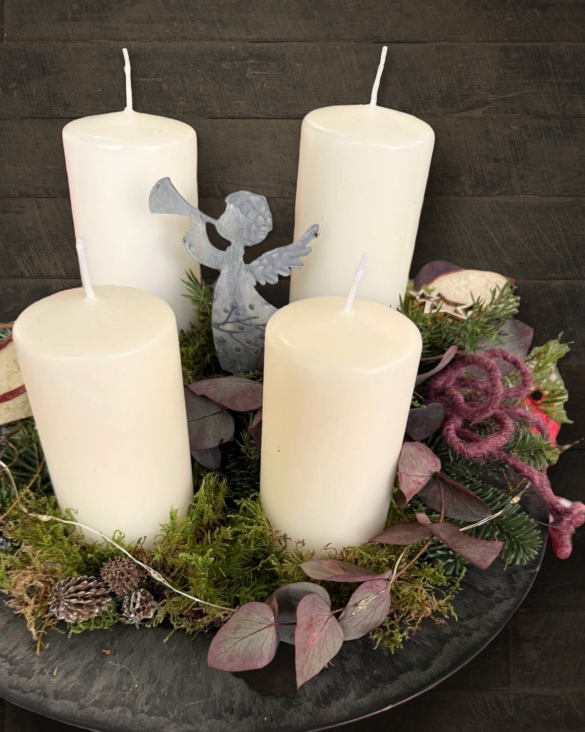 Adventsgesteck mit vier weißen Kerzen, Moos, Zapfen und Eukalyptus auf einer Schieferplatte